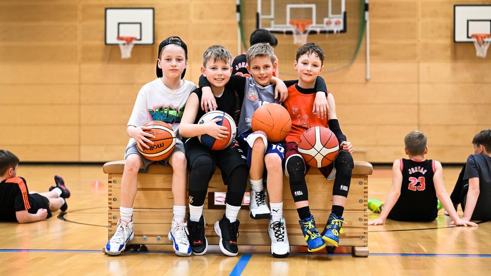 Vier Kinder sitzen auf einer Bank in der Turnhalle, halten Basketballs und lachen. Im Hintergrund sind weitere Kinder aktiv.