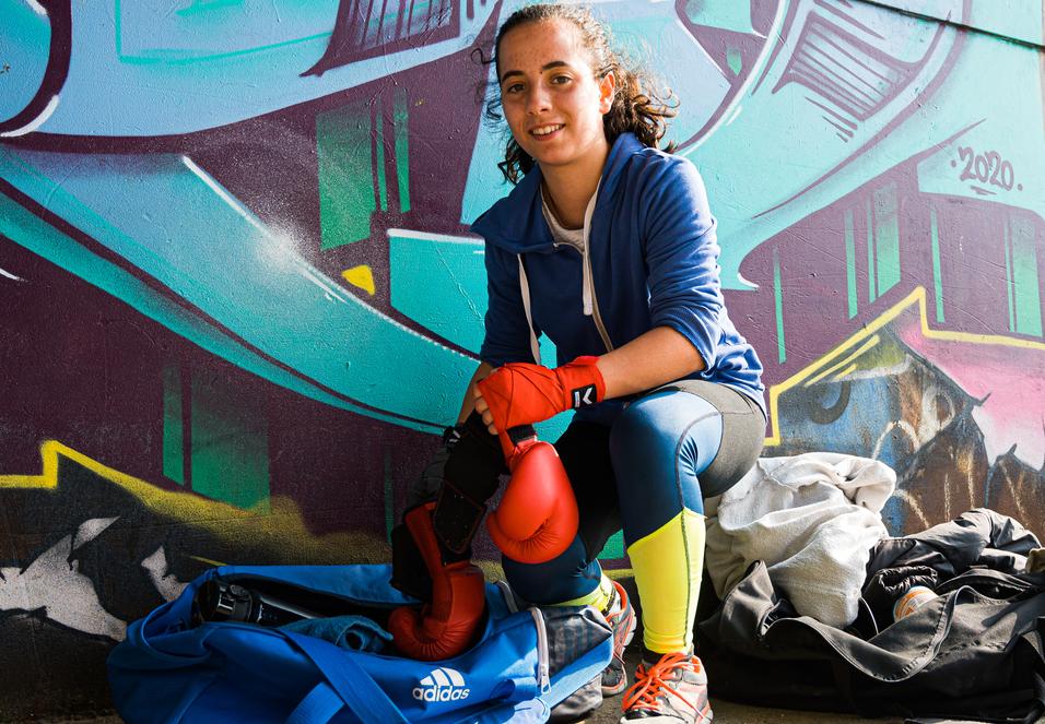 Junge Frau mit Boxhandschuhen sitzt auf dem Boden neben einer Sporttasche vor einer graffitiartigen Wand.